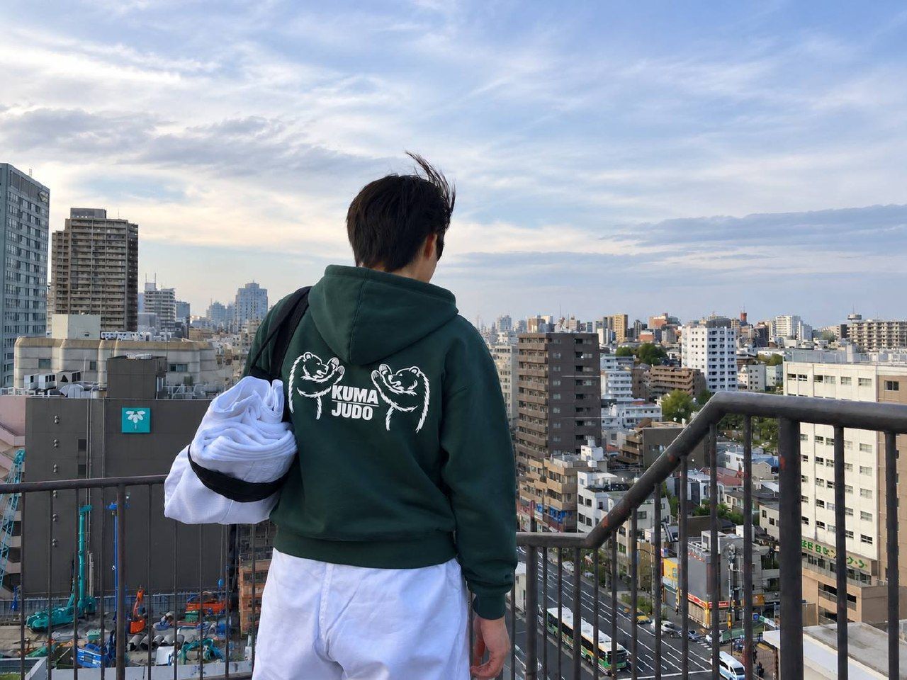A person in a KUMA JUDO hoodie with a judogi over one shoulder, on a balcony overlooking a Japanese city street