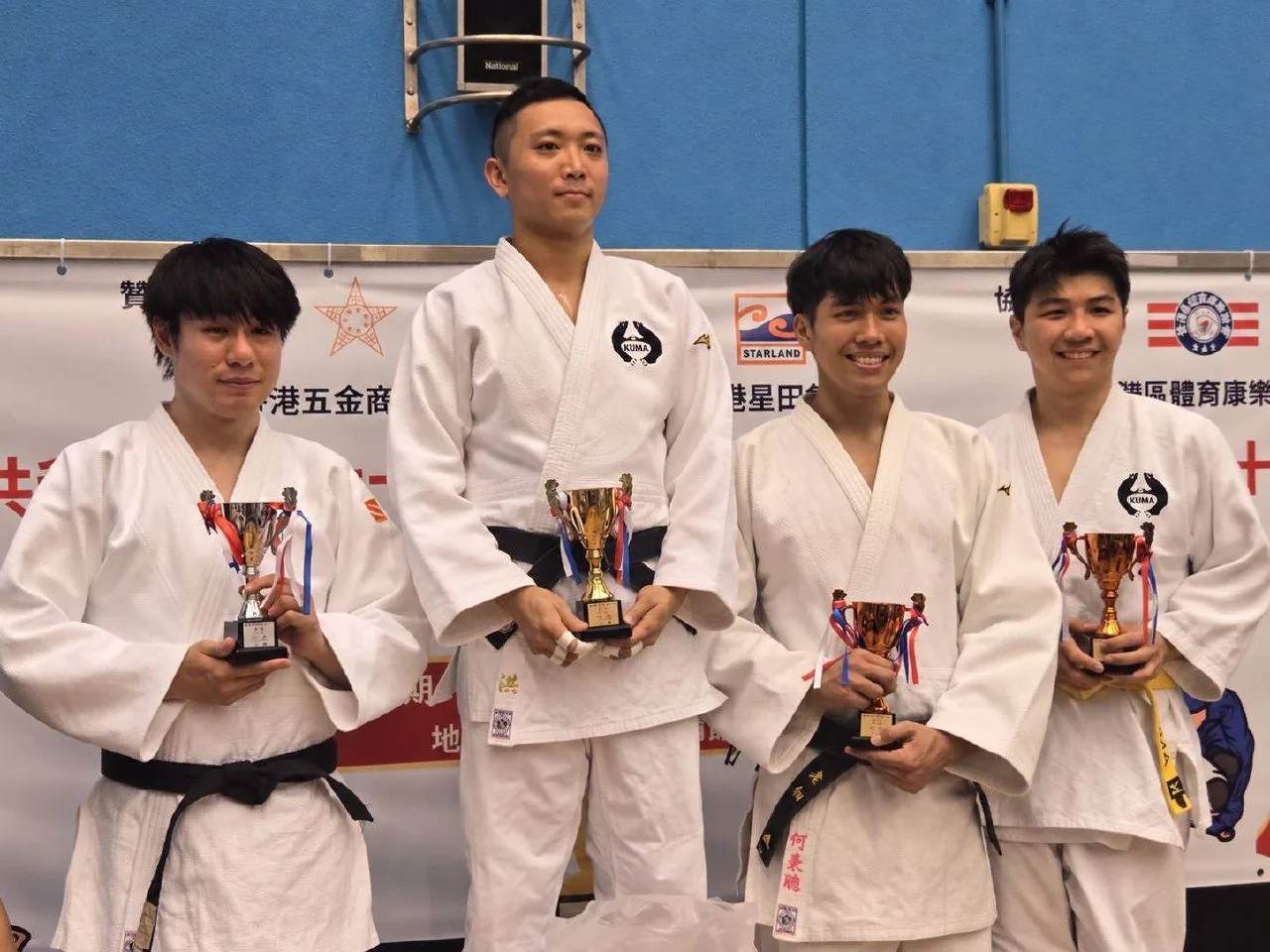 Four young judo athletes in white gis and black belts holding trophies in front of the event backdrop